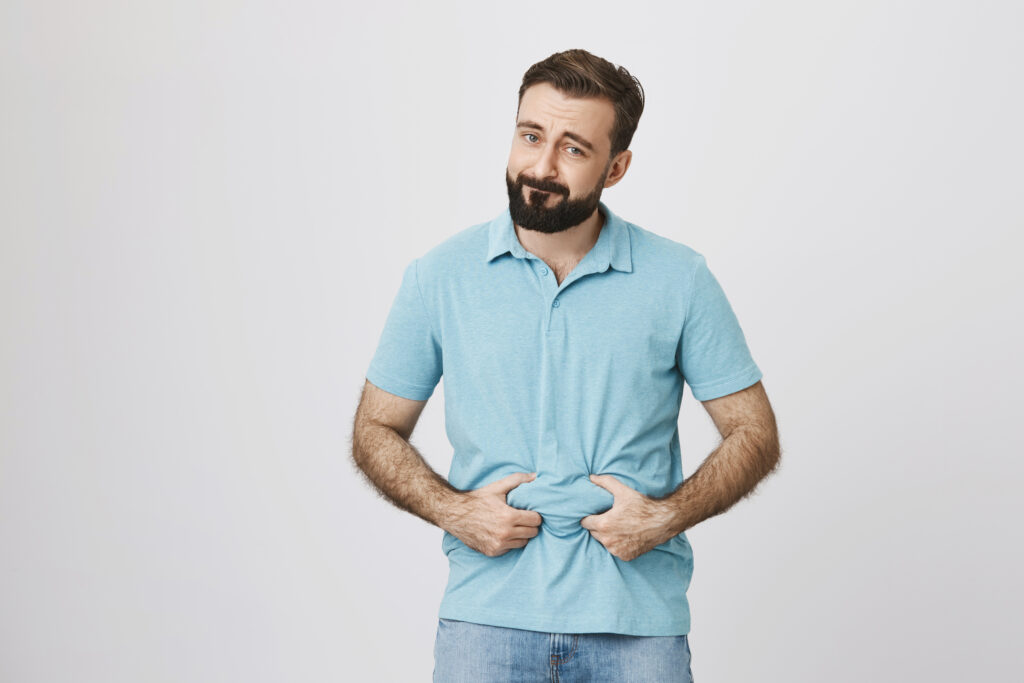 Picture of a bearded handsome man showing his beer belly over white background. Person is bothered because of his extra weight so he decides to sit on diet
